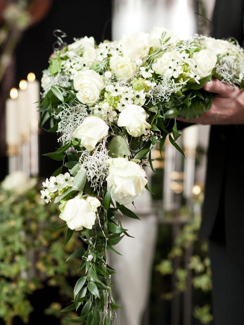 Nahaufnahme eines Blumenkranzes mit weißen Rosen, gehalten von einer Person vor einem silbernen Urnengrab.