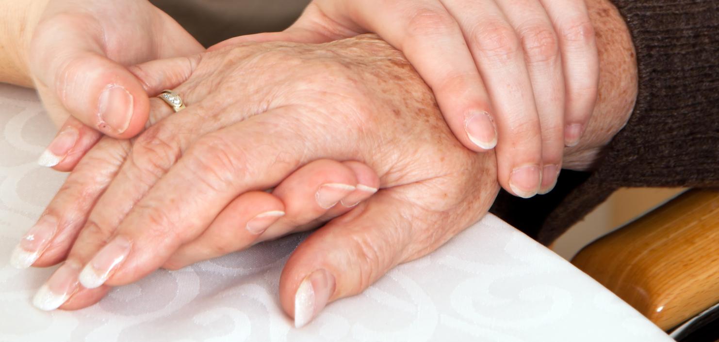 Zwei Hände, eine ältere und eine jüngere, liegen auf einem Tisch, wobei die jüngere Hand die ältere sanft umschließt.