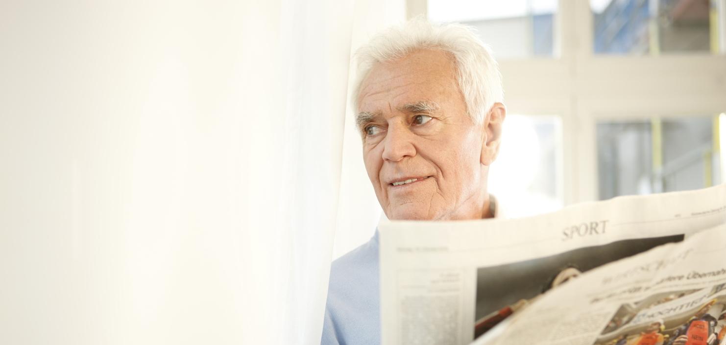 Älterer Mann mit weißem Haar liest eine aufgeschlagene Zeitung, während er an einem Fenster steht.